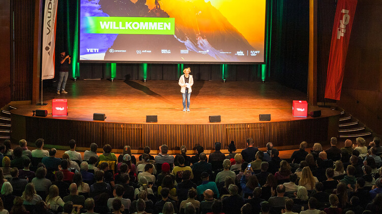 Moderatorin auf der B&uuml;hne eines Banff Tour Events vor einer Leinwand mit der Aufschrift &bdquo;Willkommen" und einem leuchtenden Bergmotiv, davor das sitzende Publikum.