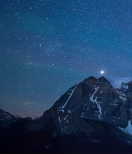 Kletterer:in an einer steil &uuml;berh&auml;ngenden, orange leuchtenden Felswand, im Hintergrund der Vollmond &uuml;ber zerkl&uuml;fteten Felst&uuml;rmen vor blauem Himmel.