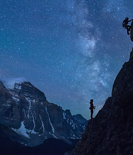 Climber on a steep, overhanging rock face glowing orange, with a full moon rising over jagged rock spires against a blue sky in the background.