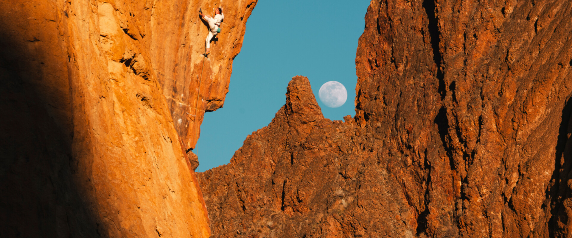 Climber on a steep, overhanging rock face glowing orange, with a full moon rising over jagged rock spires against a blue sky in the background.