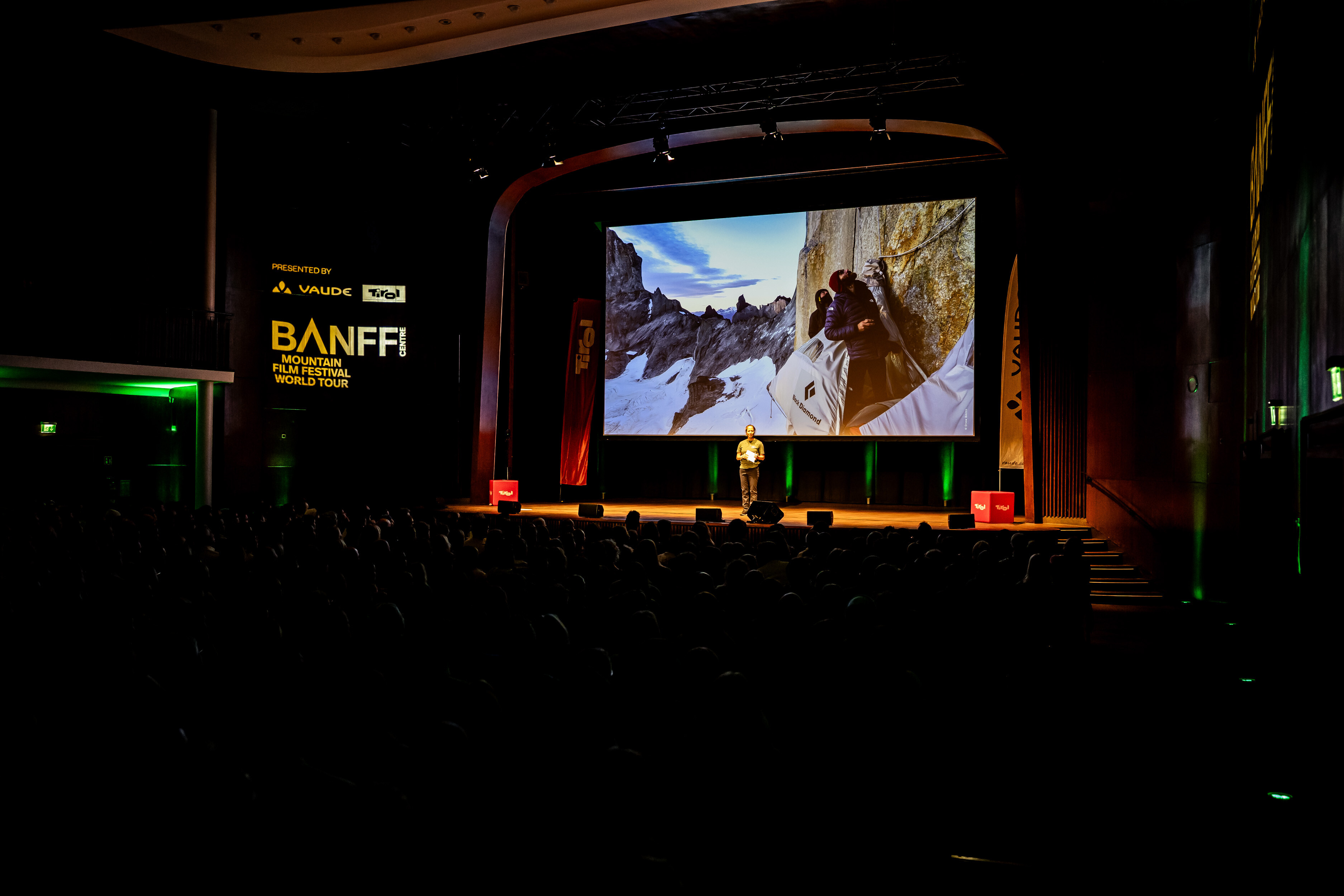 Host on stage at a Banff Tour event in front of a large screen showing two climbers in a portaledge on a rock face.