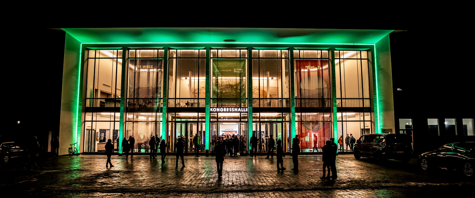 Illuminated entrance of the Alte Kongresshalle in Munich at night with visitors on the forecourt of a Banff Tour event.