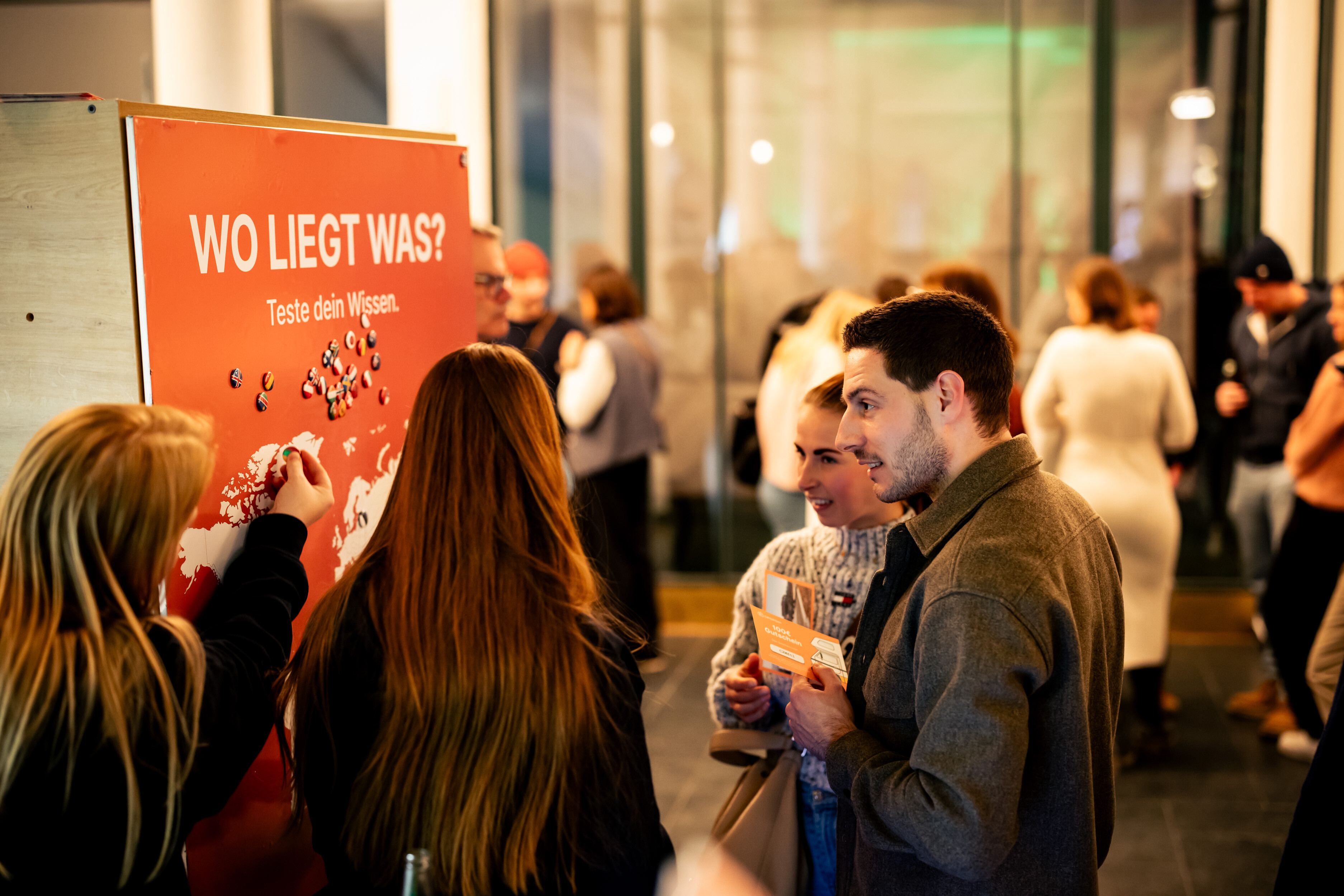 Visitors talking at a "Wo liegt was?" quiz wall in the foyer of a Banff Tour event.