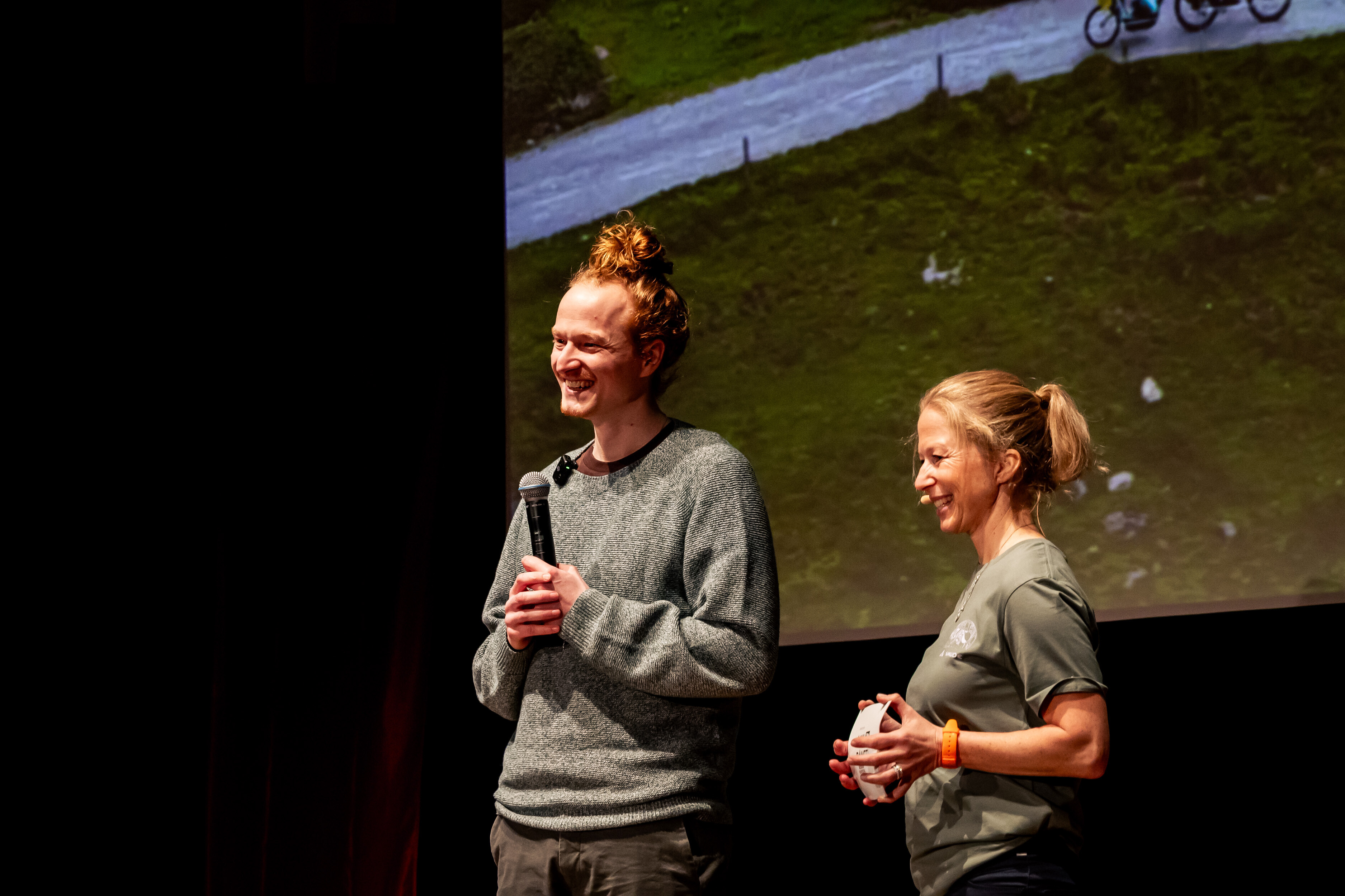 Holger Wimmer standing on stage with a microphone, laughing, at a Banff Tour event in front of a screen showing a cycling scene.