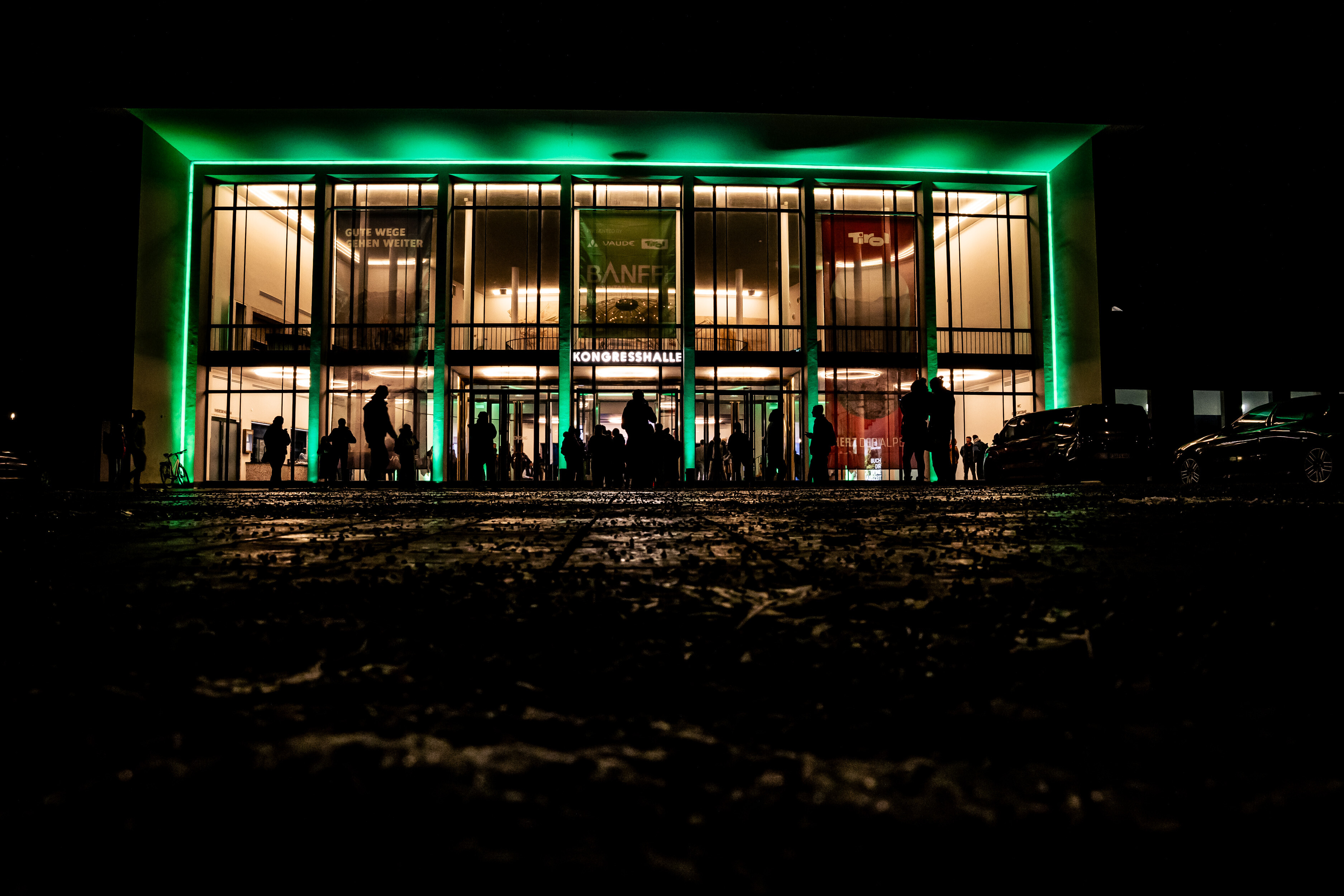 Green-lit facade of the Alte Kongresshalle in Munich at night with visitors in front of the entrance to a Banff Tour event.