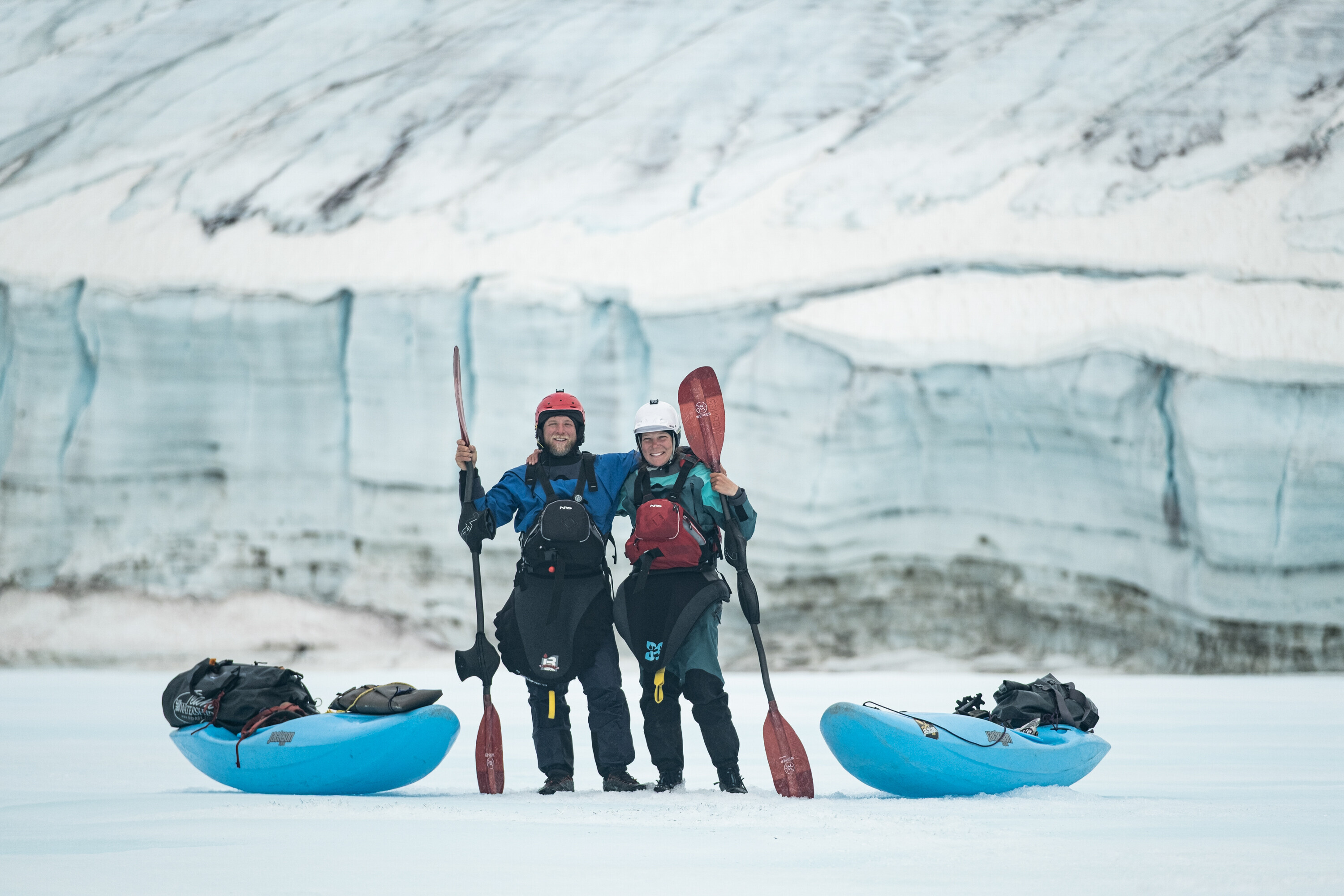 Zwei Kajakfahrer:innen, Sarah McNair-Landry and Erik Boomer, in Trockenanz&uuml;gen mit Paddeln stehen l&auml;chelnd zwischen zwei blauen Kajaks auf einem Gletscher vor einer Eiswand.